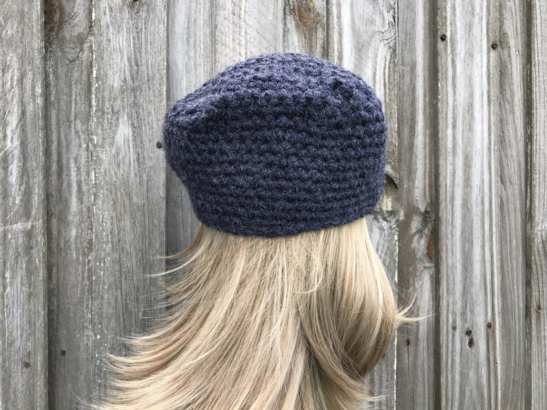 May include: A dark blue crocheted beret hat. The hat is made of a textured yarn and has a rounded crown. The beret is worn on a person with blonde hair, against a weathered wooden background.