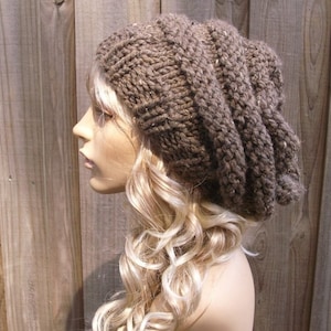 Puede incluir: Un gorro de punto marrón, holgado y caído, con un patrón de punto texturizado. El gorro se lleva en un maniquí con el pelo largo, ondulado y rubio.