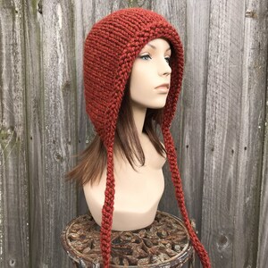 May include: A rust-colored, hand-knitted hat with ear flaps and long braided ties. The hat is displayed on a mannequin head against a weathered wooden fence. The hat's texture and color suggest warmth and a rustic aesthetic.