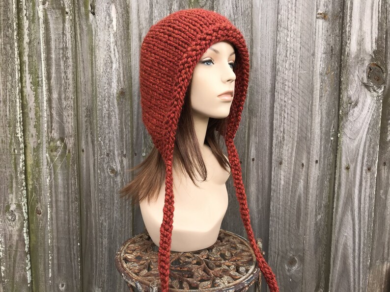 May include: A rust-colored, hand-knitted hat with ear flaps and long braided ties. The hat is displayed on a mannequin head against a weathered wooden fence. The hat's texture and color suggest warmth and a rustic aesthetic.