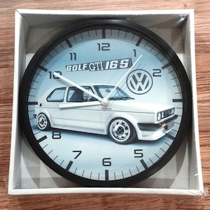 Puede incluir: Reloj blanco y negro con un coche Volkswagen Golf GTI 16S blanco en la esfera. El reloj tiene agujas negras y números negros. El coche está sobre un fondo azul claro. El reloj está en una caja blanca.