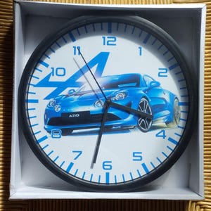 Può includere: Un orologio con cornice nera con numeri blu e un'immagine di un'auto blu e bianca. L'auto è una Alpine A110 blu con il numero 4 sul lato. Il quadrante dell'orologio ha i numeri da 1 a 12 e le parole "A110" sull'auto.