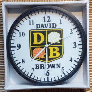 May include: A black and white clock with a black frame and white face. The clock has a black and yellow crest with the letters "D" and "B" in the centre. The crest also has a white rose and a red rose. The clock face has black numbers and hands. The clock face also has the text "DAVID BROWN" at the top.