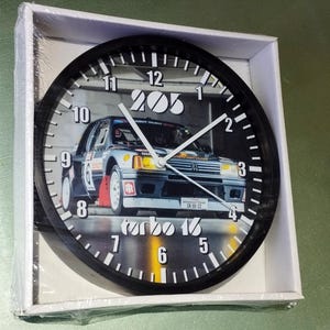 Peut inclure: Horloge murale noire avec un cadran blanc et des aiguilles noires. Le cadran de l'horloge présente une image en noir et blanc d'une Peugeot 205 Turbo 16 de rallye argentée avec le texte "205" et "turbo 16" sur le cadran.