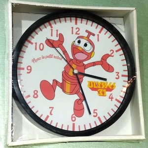 Puede incluir: Un reloj blanco y negro con un personaje de robot rojo en la cara. El robot está corriendo y tiene un cuerpo a rayas rojo y amarillo. La cara del reloj también tiene el texto "Mono le petit robot" y "ULYSSES 31".