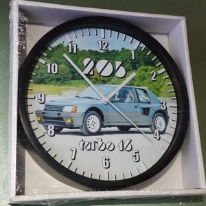Peut inclure: Une horloge murale noire et blanche avec une image d'une Peugeot 205 Turbo 16 argentée. Le cadran de l'horloge a des chiffres et des aiguilles noirs.