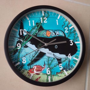 Peut inclure: Une horloge murale à cadre noir avec une scène sous-marine bleue, verte et blanche. Le cadran de l'horloge présente un requin noir et blanc avec un sous-marin sur le dessus. Les chiffres de 1 à 12 sont blancs et les aiguilles de l'horloge sont argentées.
