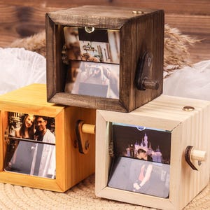 Op de afbeelding: Drie houten fotokubussen in verschillende tinten bruin en geel. Elke kubus heeft een rechthoekige opening met een foto. Een klein houten handvat is aan de zijkant van elke kubus bevestigd.