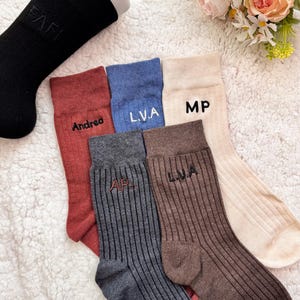 May include: A selection of ribbed socks in colours such as black, red, blue, beige, grey, and brown. Each sock is embroidered with initials or a name. The socks are arranged on a white, fluffy surface.