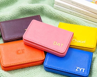 May include: Five rectangular leather card holders in purple, pink, orange, yellow, and blue. The pink holder is embossed with DAT, the yellow with Chasel, the orange with CT, and the blue with ZYT. Each case features stitched edges and a flap closure.