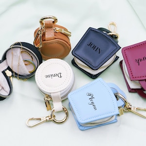 May include: A selection of personalised jewellery travel cases in different shapes and colours. Includes a black round case, a brown cylindrical case, a white round case with "Denise", a blue square case with "Megan", a navy blue square case with "John", and a burgundy square case with "Sarah".