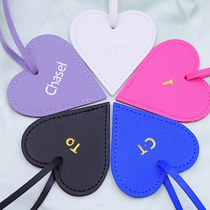 Puede incluir: Cinco llaveros de cuero en forma de corazón de diferentes colores y con nombres personalizados. Los llaveros son morado, blanco, rosa, negro y azul. Los nombres en los llaveros son "Chase", "DAT", "T", "10" y "C1".