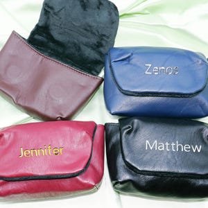 Puede incluir: Cuatro estuches de cuero personalizados en burdeos, azul, rojo y negro. Cada estuche tiene una solapa y está grabado con un nombre: "Zenos", "Jennifer" y "Matthew". El estuche burdeos está abierto, revelando un interior negro.