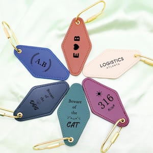 Könnte beinhalten: Eine Sammlung rautenförmiger Schlüsselanhänger in verschiedenen Farben, darunter Blau, Rosa und Lila. Jeder Schlüsselanhänger hat einen goldfarbenen Verschluss und einzigartige Texte oder Grafiken, wie Initialen, eine Sonne oder den Schriftzug "Beware of the Dog."