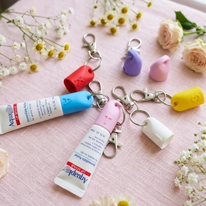 Op de afbeelding: Een verzameling lipreparatietubes met kleurrijke sleutelhangers. De tubes zijn voorzien van het label "Aquaphor LIP REPAIR" en worden vergezeld door rode, blauwe, roze, witte en gele sleutelhangeraccessoires. De afbeelding is geplaatst op een roze ondergrond met bloemaccenten.