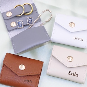 Pode incluir: Três pequenas caixas de joias retangulares de couro com fecho de pressão. As caixas são em tons de castanho, cinzento e branco. Cada caixa tem um botão dourado e um nome impresso na frente: "Angela", "Leila" e "Chasel".