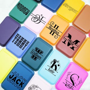 Puede incluir: Una colección de carteras para teléfonos de colores, cada una con un diseño único. Las carteras son de varios colores, incluyendo verde, rosa y azul, y presentan nombres e iniciales personalizados en letras negras. Las carteras son rectangulares y parecen ser de cuero.