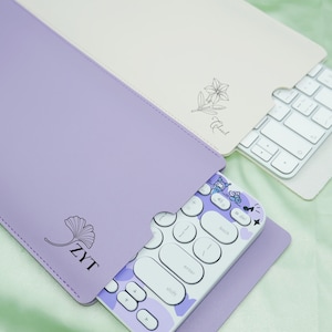 Puede incluir: Una funda de teclado de color lila claro con una hoja de ginkgo y "ZYT" impreso, que contiene un teclado blanco y morado. También se ve una funda color crema con un diseño floral y un teclado blanco. Los teclados son para un ordenador.