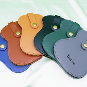May include: Five leather phone cases in different colors, including blue, brown, green, and gray. The gray case has the name "Denise" written on it.