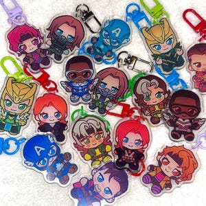 Peut inclure: Une collection de porte-clés en acrylique présentant des illustrations de style chibi de divers personnages de bandes dessinées, chacun attaché à un fermoir mousqueton en plastique coloré. Les personnages portent des costumes de super-héros.