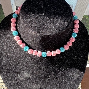 May include: A pink and blue beaded necklace with alternating colors. The beads are small and round, and they are covered in tiny crystals.