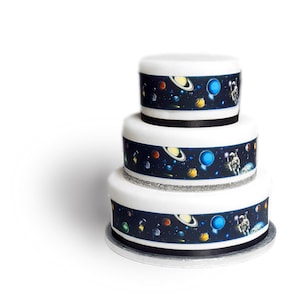 Peut inclure: Un gâteau blanc à trois étages décoré d'images comestibles de planètes, d'étoiles et d'un astronaute. Le gâteau est entouré d'un ruban noir et de paillettes argentées.