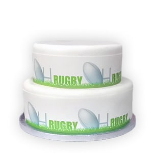 May include: A two-tiered white cake with a rugby theme. The cake is decorated with a green grass border, rugby balls, and goal posts. The word "RUGBY" is printed in green.