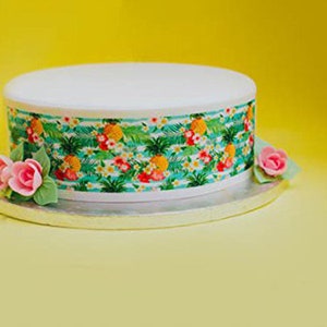 Op de afbeelding: Een ronde taart met witte glazuur en een decoratieve band met een tropisch bloemenpatroon met ananassen en bloemen. Roze rozen en groene bladeren sieren de basis, tegen een gele achtergrond.