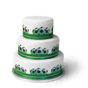 May include: A three-tiered white cake decorated with green grass and footballs. The cake is on a silver cake stand.