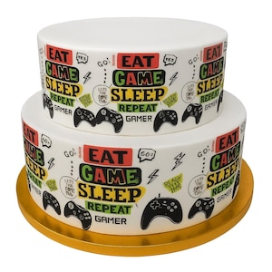 May include: A two-tiered white cake with a gold base, decorated with the words "EAT GAME SLEEP REPEAT GAMER" and controller graphics. The cake has a playful design with colorful text and illustrations.