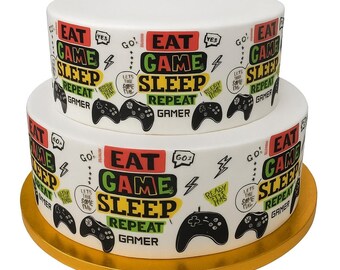 May include: A two-tiered white cake with a gold base, decorated with the words "EAT GAME SLEEP REPEAT GAMER" and controller graphics. The cake has a playful design with colorful text and illustrations.