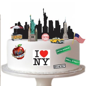 May include: A white cake decorated with a New York City theme. The cake features a black silhouette of the New York City skyline, a Statue of Liberty figurine, a yellow taxi cab, a red apple with "I ♥ NY" and "The Big Apple" text, and a red, white, and blue American flag.