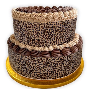 May include: A two-tiered cake decorated with brown and beige frosting and a leopard print design. The cake is on a gold cake board.