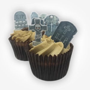 Może przedstawiać: Dwie babeczki w brązowych papilotach z jasnobrązowym kremem. Każda babeczka jest ozdobiona topperem w kształcie nagrobka z napisem "R.I.P." i motywem czaszki. Motyw Halloween.