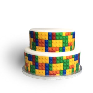 Puede incluir: Un pastel de dos pisos decorado con un patrón de bloques de Lego de colores. El pastel es blanco con un patrón de bloques de Lego amarillos, rojos, azules y verdes alrededor del piso inferior y el piso superior.