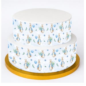 May include: Two-tiered cake with white icing and a Peter Rabbit design. The cake's sides feature a repeating pattern of Peter Rabbit holding flowers and balloons on a light blue background. The cake sits on a gold-coloured base.