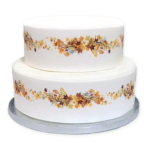 Può includere: Torta bianca a due piani decorata con un bordo di foglie autunnali marroni, arancioni e gialle. La torta è su un supporto per torte argentato.