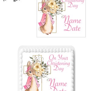May include: A 7.5 inch square edible icing topper featuring a watercolour illustration of a bunny rabbit in a pink jacket standing in front of a gold cross with white flowers. The text "On Your Christening Day" and "Name Date" is printed in pink.
