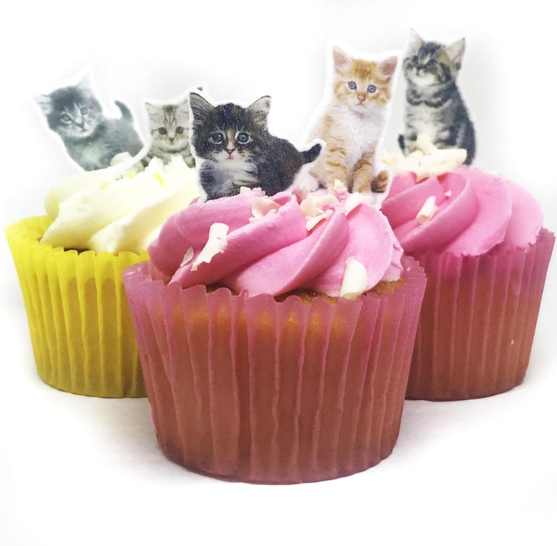 May include: Three cupcakes with yellow and pink liners, topped with white and pink frosting. Each cupcake is decorated with a kitten-shaped cutout. The cupcakes are on a white background.