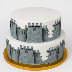 May include: A two-tiered cake with white frosting and a gray castle design. The castle walls are detailed with stone textures, windows, and battlements. The cake sits on a gold-colored cake board, perfect for a themed celebration.