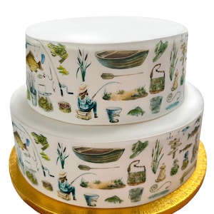 May include: A two-tiered white cake decorated with a white icing and a printed edible image of a fishing scene. The image features a person fishing, a boat, fishing poles, frogs, and other fishing-related items. The cake is on a gold base.