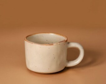 Peut inclure: Une tasse en céramique crème avec une anse arrondie. La tasse a une texture tachetée et un bord légèrement irrégulier avec un bord marron. La tasse est posée sur un fond marron uni.