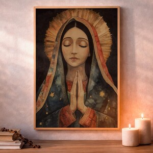 Light wood frame hanging on a pale textured wall above a wooden tabletop, showing a watercolor of Our Lady of Guadalupe with eyes closed and hands clasped in prayer, wearing a blue starry mantle with red trim and a radiating golden halo on a dark background. A stack of books with a rosary lies at left, and two lit white pillar candles sit at right in warm light.
