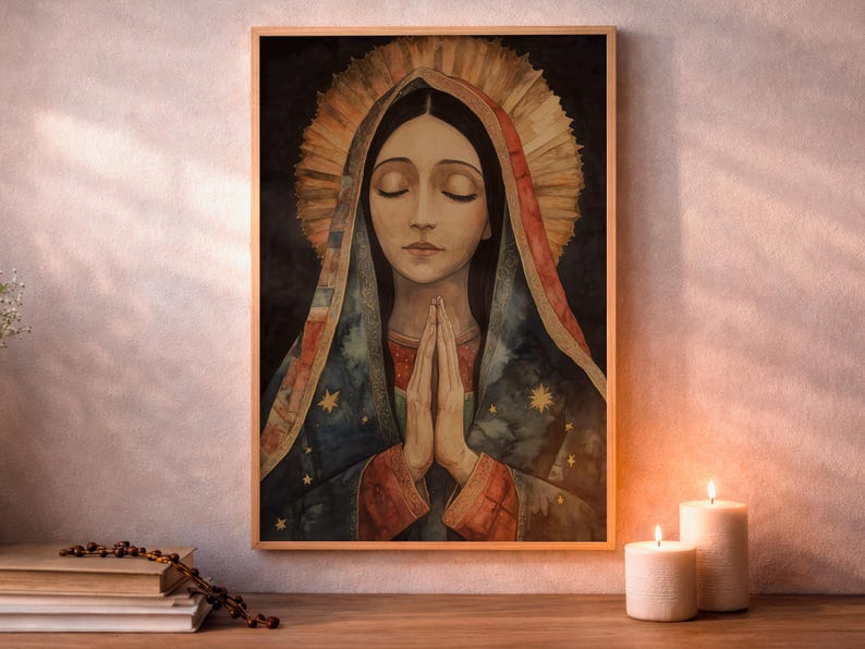 Light wood frame hanging on a pale textured wall above a wooden tabletop, showing a watercolor of Our Lady of Guadalupe with eyes closed and hands clasped in prayer, wearing a blue starry mantle with red trim and a radiating golden halo on a dark background. A stack of books with a rosary lies at left, and two lit white pillar candles sit at right in warm light.