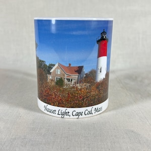 May include: A cylindrical mug featuring a scenic image of Nauset Light, Cape Cod, Mass. The image includes a red and white lighthouse, a house with a red roof, and autumn foliage under a blue sky. The text "Nauset Light, Cape Cod, Mass" is printed on the bottom.
