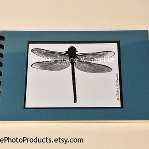 May include: A spiral-bound notebook with a teal cover. The notebook features a black and white dragonfly illustration on a white background, framed by a black border. The text "© 2015 Duane V. Gamble" is visible.
