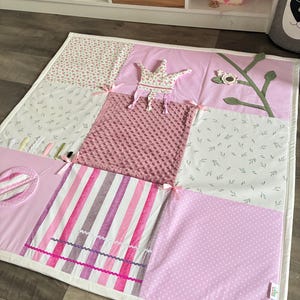 May include: A patchwork baby play mat in shades of pink and white. The mat features various patterns, including floral prints, stripes, and polka dots. A fabric crown and a branch with a flower are sewn onto the mat. The mat is trimmed with a white border.