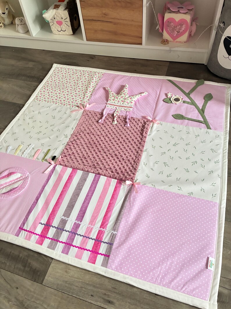 May include: A patchwork baby play mat in shades of pink and white. The mat features various patterns, including floral prints, stripes, and polka dots. A fabric crown and a branch with a flower are sewn onto the mat. The mat is trimmed with a white border.