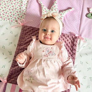 May include: A baby wearing a pink dress with floral embroidery and matching leggings. The baby is wearing a headband with a crown design. The baby is lying on a patchwork quilt with floral and polka dot patterns.
