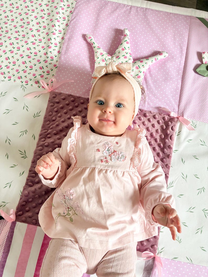 May include: A baby wearing a pink dress with floral embroidery and matching leggings. The baby is wearing a headband with a crown design. The baby is lying on a patchwork quilt with floral and polka dot patterns.